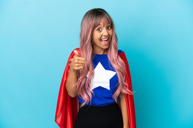 Mulher jovem super-heroína com cabelo rosa isolado em um fundo azul e polegar para cima porque algo bom aconteceu