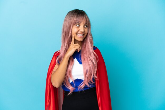 Mulher jovem super-heroína com cabelo rosa isolado em um fundo azul tendo uma ideia enquanto olha para cima