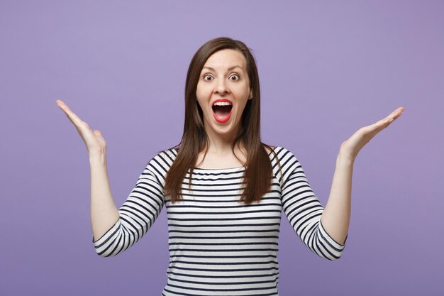 Mulher jovem surpresa chocada em roupas listradas casuais posando isolado no fundo roxo violeta no estúdio. Conceito de estilo de vida de pessoas. Mock up espaço de cópia. Mantendo a boca bem aberta, espalhando as mãos.