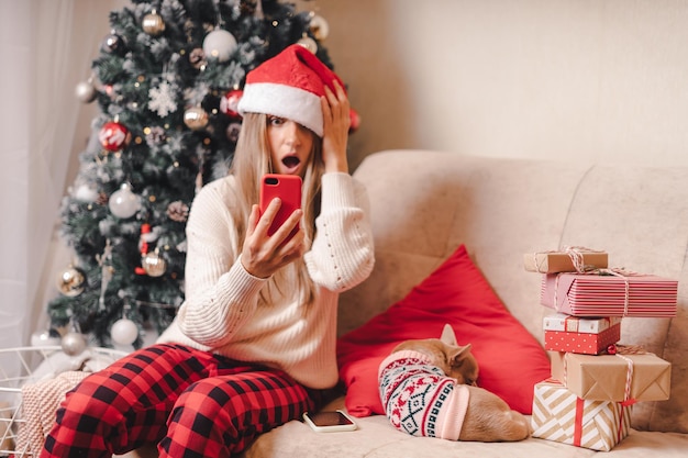 Mulher jovem surpresa e cachorro vestindo suéter tira uma selfie no smartphone nas férias de inverno