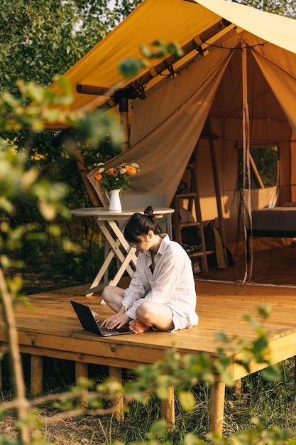 Mulher jovem trabalha em laptop enquanto está sentada relaxada em uma cadeira ao lado da fogueira viajando com tenda Conceito de trabalho remoto e fuga para a natureza