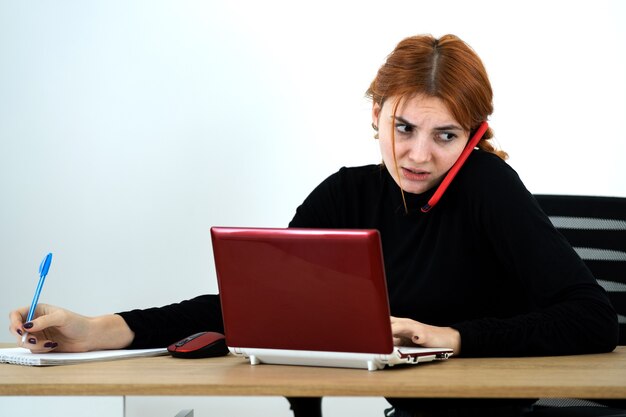 Mulher jovem trabalhador de escritório falando em um telefone celular, sentado atrás da mesa de trabalho com o laptop e o notebook.