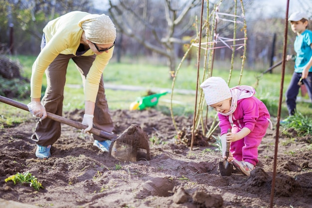 Mulher jovem trabalhando no jardim enquanto sua filha brinca com pá ao lado dela e do filho