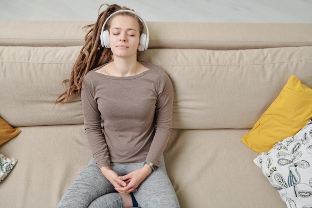 Mulher jovem tranquila com fones de ouvido ouvindo música para meditação no sofá, se divertindo