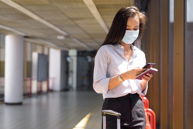 Mulher jovem turista asiática com máscara usando telefone e segurando o passaporte no aeroporto