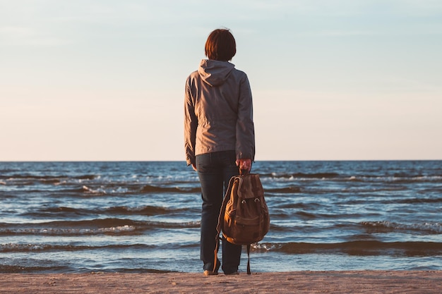 Mulher jovem turista com mochila olhando para o mar ao pôr do sol