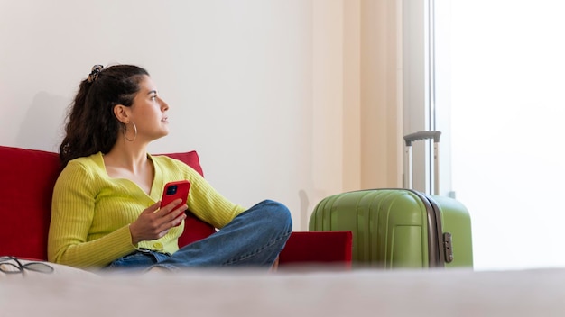 Mulher jovem turista com seu smartphone em um quarto de hotel sentado em um sofá olhando pela janela