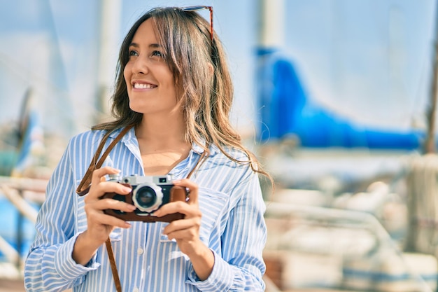 Mulher jovem turista hispânica sorrindo feliz usando câmera vintage no porto