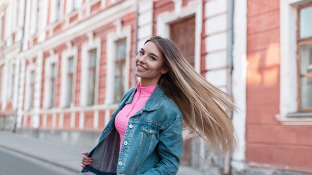 Mulher jovem urbana alegre com um sorriso fofo com cabelo comprido loiro em uma jaqueta jeans vintage em um top rosa na moda posa perto de um prédio na rua. Garota muito feliz caminha pela cidade no dia de verão.