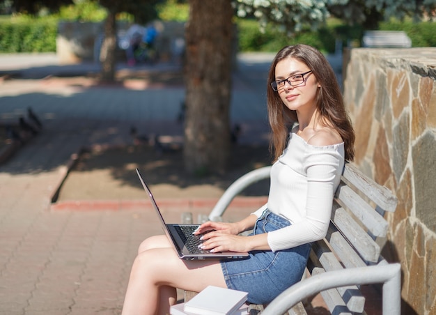 Mulher jovem usa laptop sentado em um banco do parque.