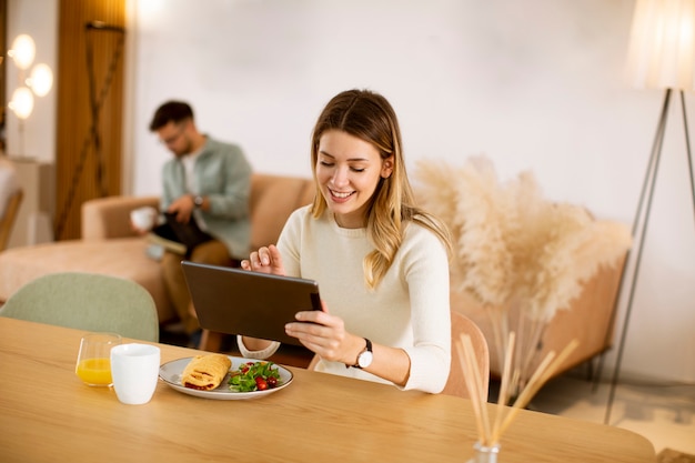 Mulher jovem usando tablet digital e tomando café da manhã na cozinha com um namorado sentado