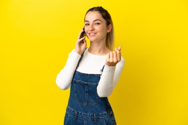 Mulher jovem usando telefone celular sobre fundo amarelo isolado fazendo gesto de dinheiro