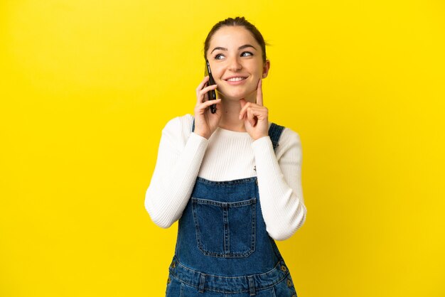 Mulher jovem usando telefone celular sobre fundo amarelo isolado, pensando em uma ideia enquanto olha para cima