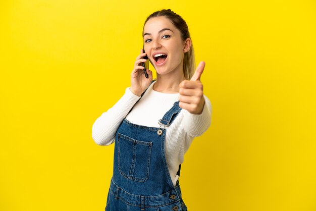 Mulher jovem usando telefone celular sobre um fundo amarelo isolado com polegar para cima porque algo bom aconteceu