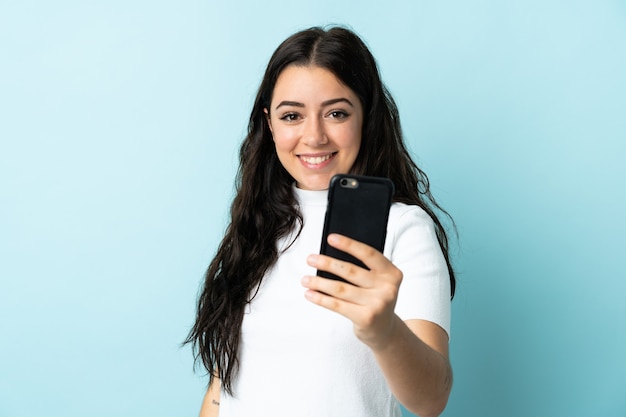 Mulher jovem usando um celular isolado em uma parede azul com uma expressão feliz