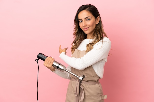 Mulher jovem usando um liquidificador manual sobre um fundo rosa isolado apontando para trás