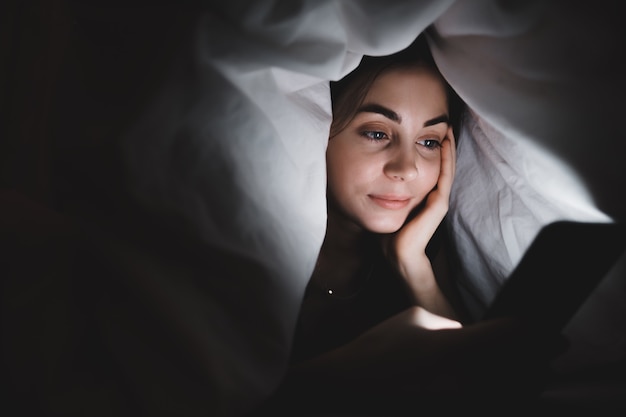 Mulher jovem usando um smartphone embaixo de um cobertor na cama