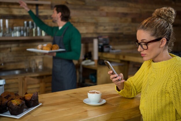 Mulher jovem usando um telefone celular enquanto um barista trabalhava em segundo plano em uma cafeteria