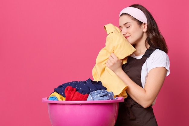 Mulher jovem veste camiseta, avental marrom e faixa de cabelo, em pé com uma bacia rosa com linho limpo, cheira roupas frescas, estando satisfeita com o trabalho realizado, fica encostada na parede rosada.