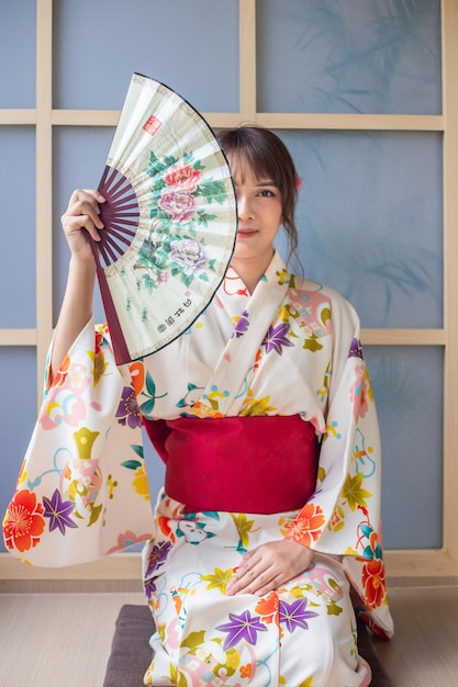 Foto mulher jovem vestindo kimono tradicional japonês segurando um ventilador de papel sentada em uma casa de estilo japonês
