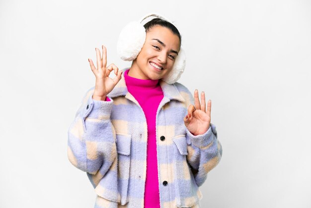 Mulher jovem vestindo muffs de inverno sobre fundo branco isolado mostrando um sinal ok com os dedos