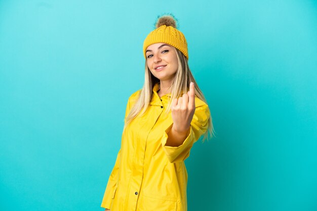 Mulher jovem vestindo um casaco à prova de chuva sobre um fundo azul isolado fazendo o gesto de aproximar-se