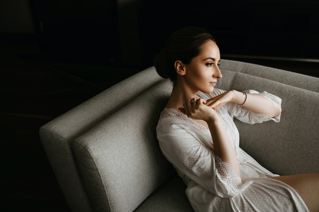 Mulher jovem vestindo um roupão de seda branco, sentada em um sofá. Humor calmo.