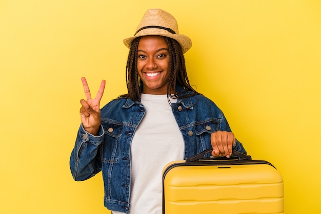 Mulher jovem viajante afro-americano segurando mala isolada em fundo amarelo, mostrando o número dois com os dedos.