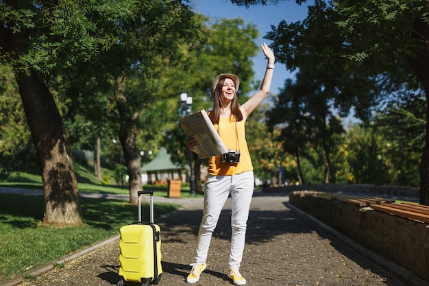 Mulher jovem viajante alegre turista com chapéu com mapa da cidade mala acenando a mão para saudação encontrar amigo na cidade ao ar livre. garota viajando para o exterior para viajar no fim de semana. estilo de vida da viagem de turismo.