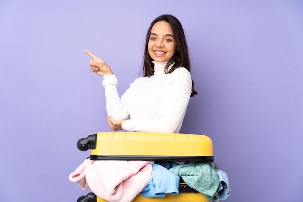 Mulher jovem viajante com uma mala cheia de roupas sobre um fundo roxo isolado apontando o dedo para o lado