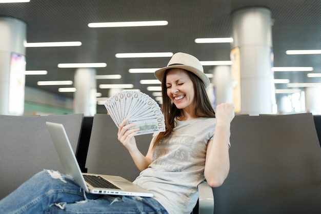 Mulher jovem viajante feliz com laptop segurando um pacote de dólares em dinheiro, fazendo o gesto de vencedor esperar no saguão do aeroporto