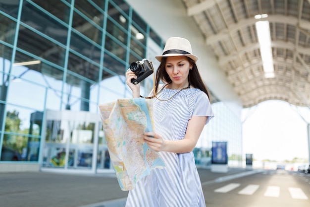 Mulher jovem viajante preocupada com um chapéu segurando uma câmera fotográfica vintage retrô, olhando para o mapa de papel no aeroporto internacional