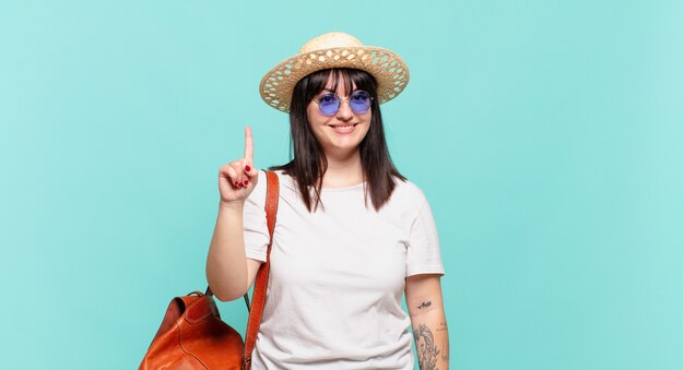 Mulher jovem viajante sorrindo e parecendo amigável, mostrando o número um ou primeiro com a mão em contagem regressiva