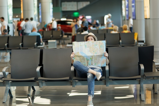 Mulher jovem viajante turista cobrindo com mapa de papel, procurando rota, esperando no saguão do aeroporto internacional