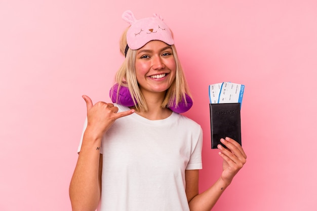 Mulher jovem viajante venezuelano segurando um passaporte isolado na parede rosa, mostrando um gesto de chamada de telefone móvel com os dedos.