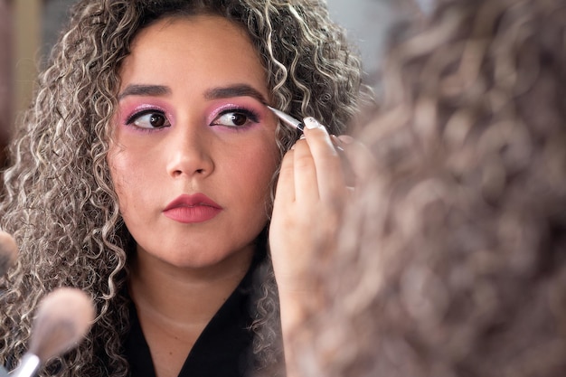 Foto mulher latina com cabelos cacheados aplicando maquiagem de sobrancelha na frente de um espelho