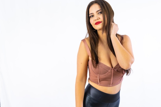 Mulher latina de aparência sedutora com cabelo preto vestindo uma blusa marrom e calça preta de látex fotografada em um estúdio com fundo branco