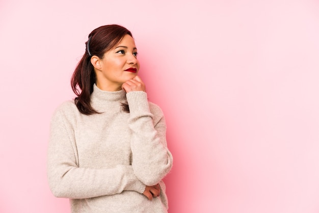 Mulher latina de meia-idade isolada em um fundo rosa, olhando de soslaio com expressão duvidosa e cética.