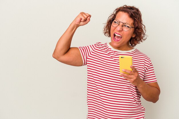 Mulher latina de meia-idade isolada no fundo branco, levantando o punho após uma vitória, o conceito de vencedor.