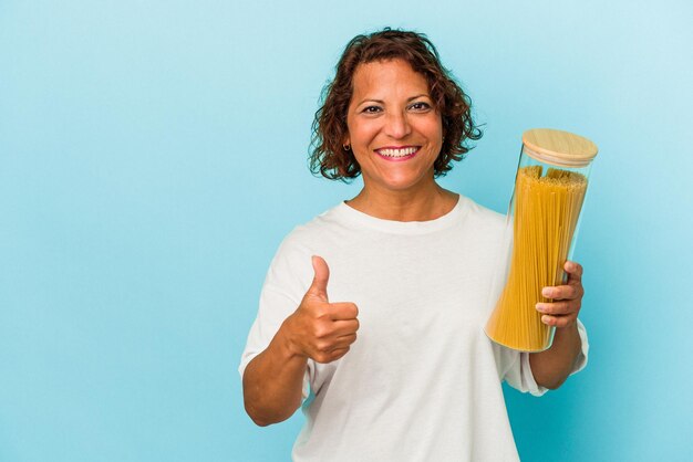Mulher latina de meia-idade segurando o pote de macarrão isolado no fundo azul, sorrindo e levantando o polegar
