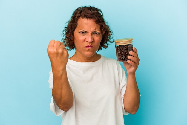 Mulher latina de meia-idade segurando uma jarra de café isolada sobre fundo azul, mostrando o punho para a câmera, expressão facial agressiva.