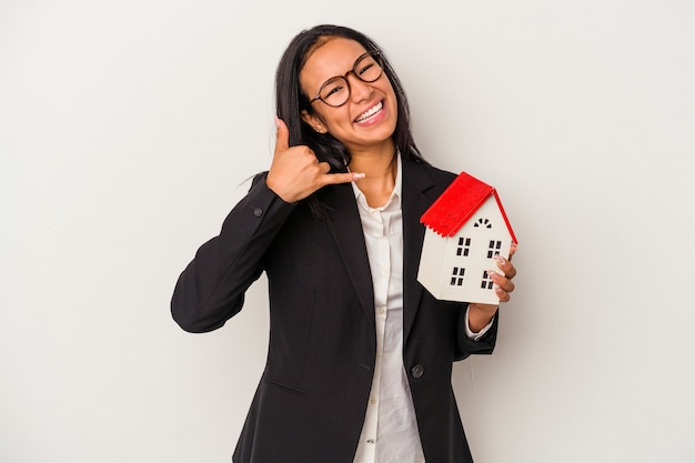 Mulher latina de negócios jovem segurando uma casa de brinquedo isolada no fundo branco, mostrando um gesto de chamada de telefone móvel com os dedos.