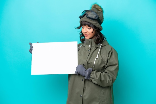 Mulher latina esquiadora com óculos de snowboard isolados em fundo azul segurando um cartaz vazio com expressão feliz