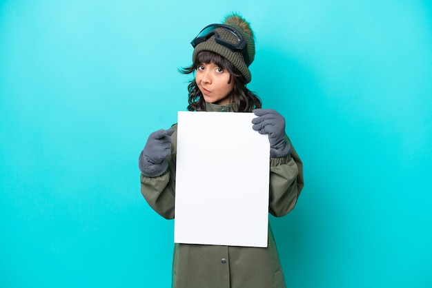 Mulher latina esquiadora com óculos de snowboard isolados em fundo azul segurando um cartaz vazio e apontando para a frente