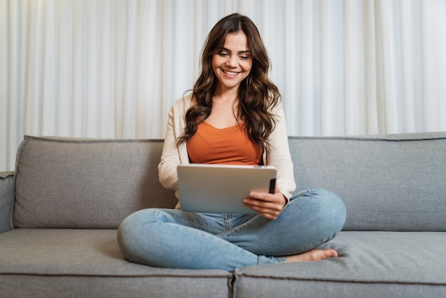 Mulher latina feliz relaxando em casa sozinha, sentada na sala, compartilhando boas notícias nas redes sociais via tablet digital. Mulher sorridente desfrutar de comida de bens de pedido de fim de semana online no aplicativo para tablet.