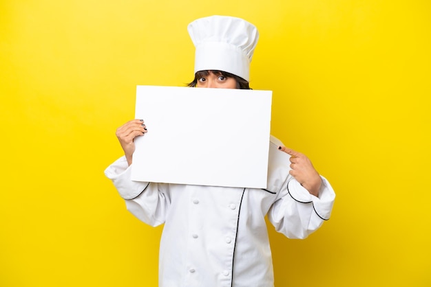 Mulher latina jovem chef isolada em fundo amarelo segurando um cartaz vazio e se escondendo atrás dele