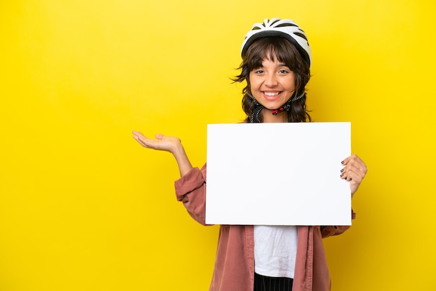 Mulher latina jovem ciclista isolada em fundo amarelo segurando um cartaz vazio com expressão de surpresa