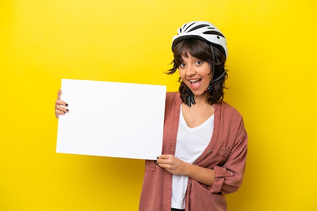 Mulher latina jovem ciclista isolada em fundo amarelo segurando um cartaz vazio com expressão feliz