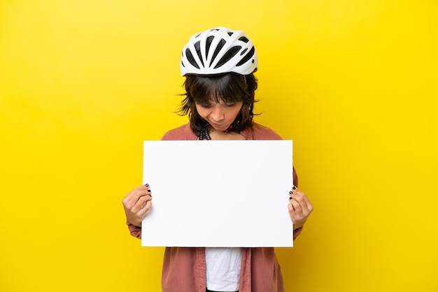 Mulher latina jovem ciclista isolada em fundo amarelo segurando um cartaz vazio e olhando para ele