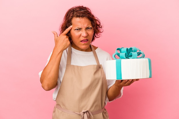 Mulher latina pastelaria de meia idade segurando um bolo isolado no fundo rosa, mostrando um gesto de decepção com o dedo indicador.